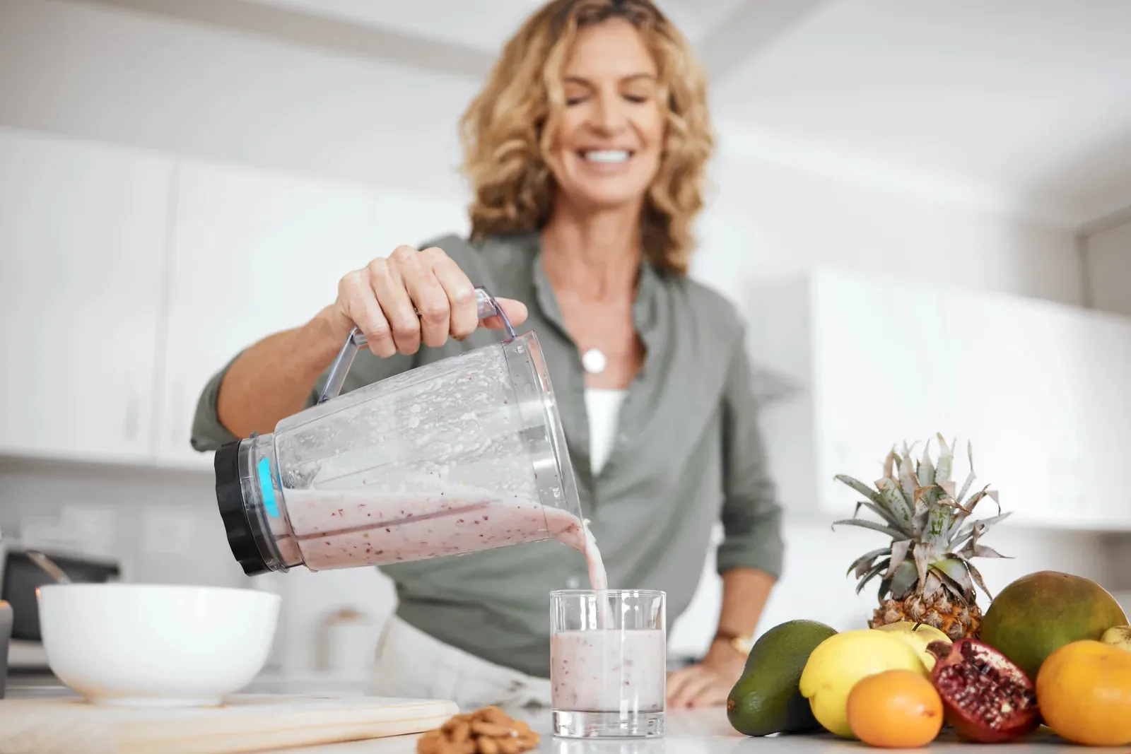 A woman pouring a smoothie from a blender jug in a kitchen, fresh fruit on the bench beside her