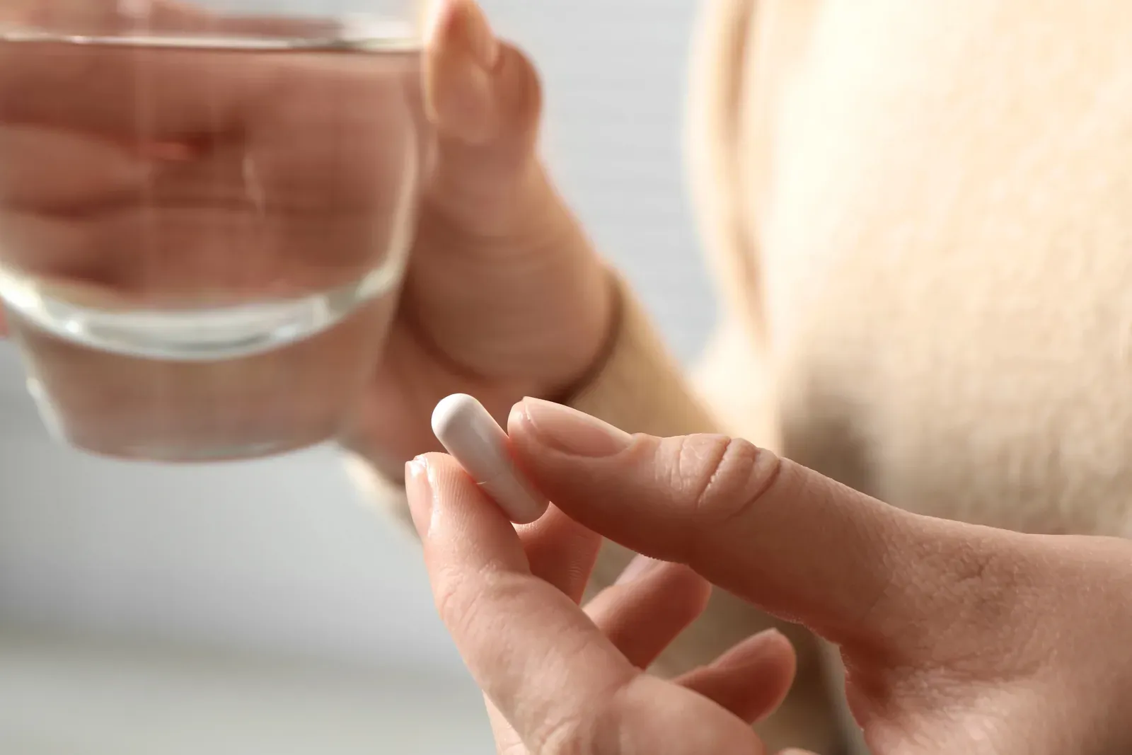A hand holding a glass of water and a single white capsule