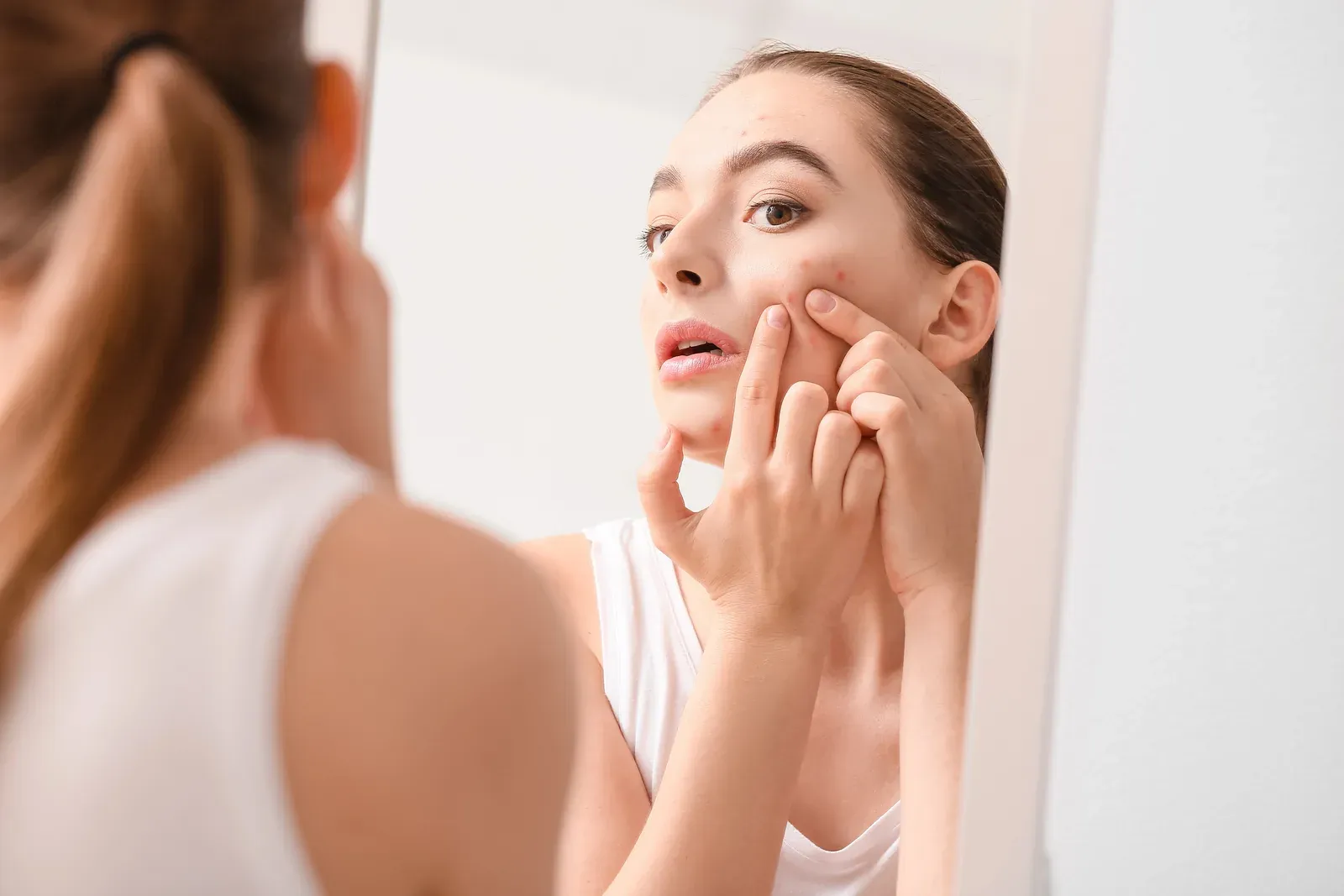 A young woman examining her cheek in a bathroom mirror